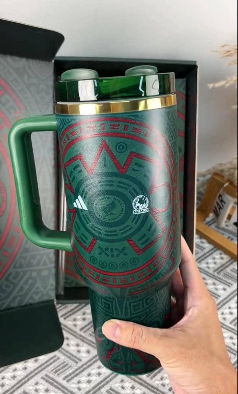 Puede incluir: Vaso t&eacute;rmico verde y rojo con asa, con un dise&ntilde;o geom&eacute;trico y un borde dorado. El vaso tiene una tapa y un asa verdes, y el logotipo "NAMLO" es visible. El vaso est&aacute; en una caja negra.