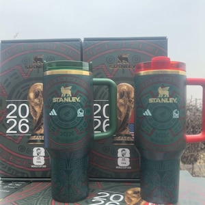 Puede incluir: Dos tazas de viaje Stanley verdes y rojas con asas, con un dise&ntilde;o decorativo y el texto "2026". Las tazas forman parte de una colecci&oacute;n FIFA World Cup 2026, con el logotipo de Adidas y el logotipo de Stanley visibles.