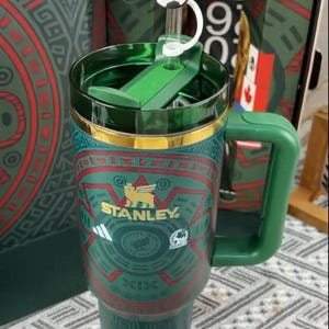 Puede incluir: Un termo de viaje Stanley verde y dorado con un adorno de tapa en forma de bal&oacute;n de f&uacute;tbol. El termo tiene un asa y un patr&oacute;n decorativo con el logotipo de Stanley. El termo mide aproximadamente 23 cm de alto.