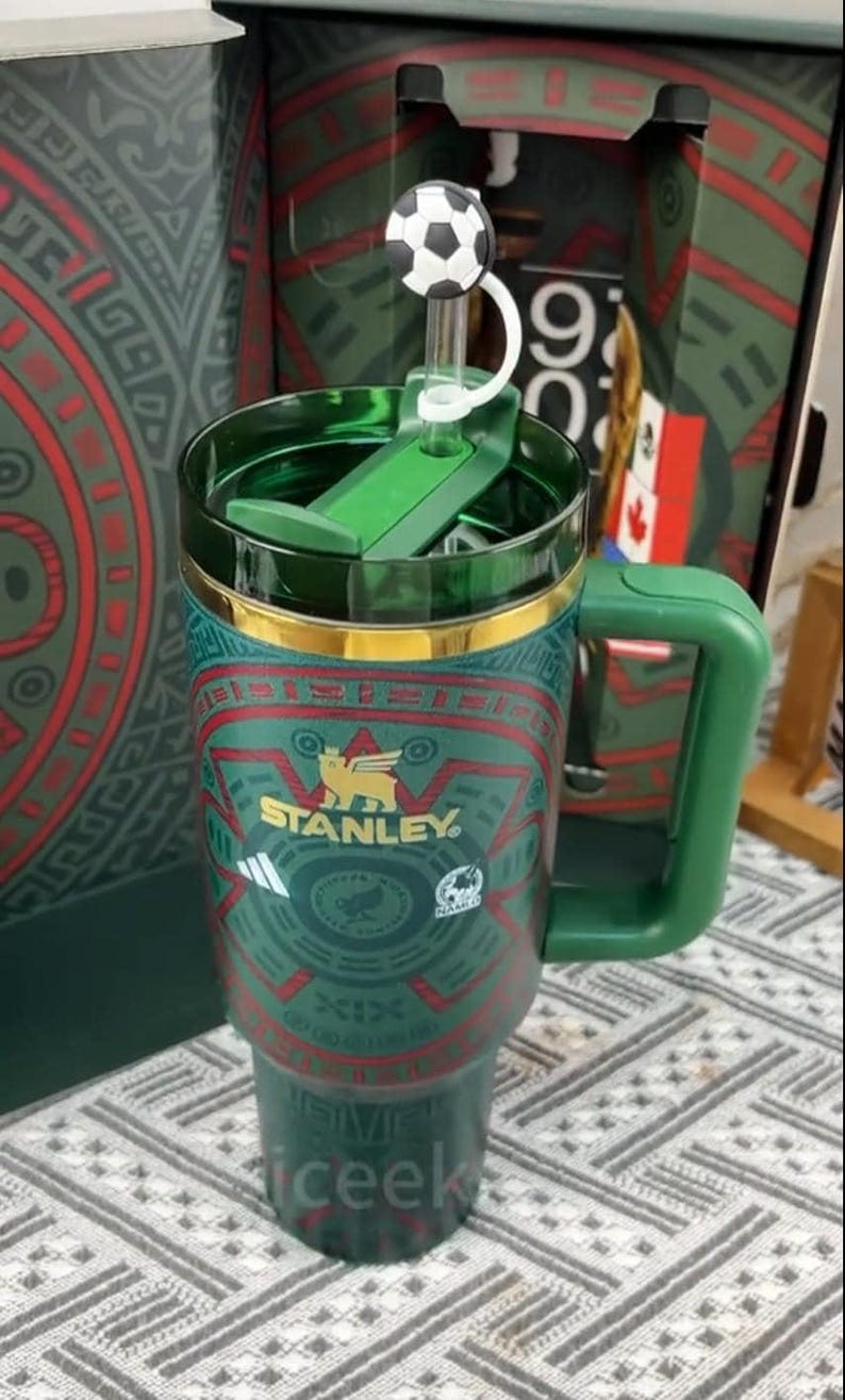 Puede incluir: Un termo de viaje Stanley verde y dorado con un adorno de tapa en forma de bal&oacute;n de f&uacute;tbol. El termo tiene un asa y un patr&oacute;n decorativo con el logotipo de Stanley. El termo mide aproximadamente 23 cm de alto.