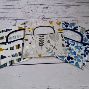 May include: Three fabric garment bags with metal hangers. The bags feature different patterns: fish, floral, and botanical designs. Each bag has a circular opening and a dark blue trim. The bags are made of fabric and have a button closure.