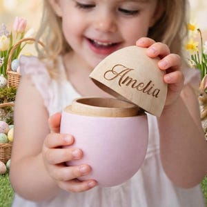 May include: A light pink, egg-shaped wooden box with a natural wood lid. The lid is engraved with the name "Amelia" in cursive. The box is being opened, revealing the interior. Easter-themed background.