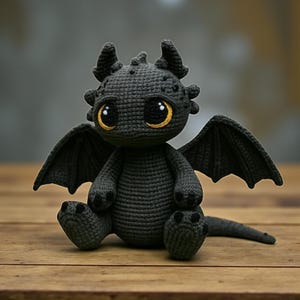 以下が含まれることがあります： 手作りの、かぎ針編みのドラゴンのぬいぐるみ。色は濃いグレー。 ドラゴンは、黄色い虹彩を持つ大きくて丸い目、小さな角、そして広げられた翼を持っています。 木の表面に座っています。