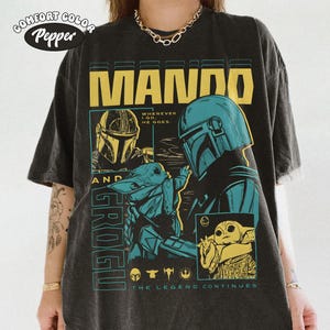 K&ouml;nnte beinhalten: Dunkelgraues Comfort Colors T-Shirt mit einem grafischen Design mit dem Text "MANDO" in Gelb. Das Design enth&auml;lt Illustrationen eines Mandalorianer-Helms und Grogu, mit dem Text "THE LEGEND CONTINUES."