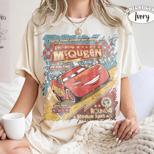 May include: Ivory-colored t-shirt featuring a graphic of Lightning McQueen from the Disney Pixar Cars movie. The design includes text that reads "Lightning McQueen" and "Radiator Springs Saturday Night Special". The shirt is a Comfort Color.