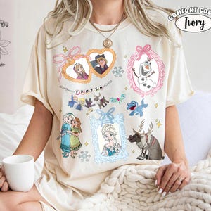 Könnte beinhalten: Elfenbeinfarbenes T-Shirt mit verschiedenen Charakteren und Elementen aus dem Animationsfilm "Die Eiskönigin". Das Design umfasst gerahmte Porträts von Charakteren, Schneeflocken und das Wort "FROZEN". Das Shirt ist von der Marke Comfort Colors.