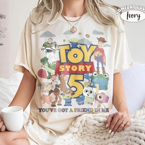 May include: An ivory t-shirt featuring a colorful graphic print of Toy Story characters including Woody, Buzz Lightyear, and Rex. The text reads Toy Story 5 and You've Got A Friend In Me. A small circular label in the corner says Comfort Color Ivory.