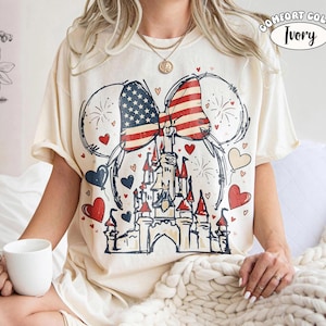 May include: Ivory Comfort Colors t-shirt featuring a patriotic design. The graphic includes a castle, a bow with the American flag, Mickey Mouse ears, hearts, and fireworks. The shirt is short-sleeved and has the words "Comfort Color Ivory" printed on it.