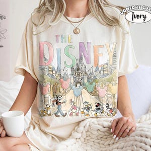 Pu&ograve; includere: T-shirt color avorio con una grafica colorata di personaggi Disney e la scritta "DISNEY". Il design include palloncini di Topolino e un castello. La maglietta &egrave; del marchio Comfort Color.