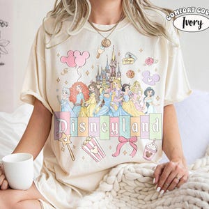 Puede incluir: Camiseta de color marfil con un dise&ntilde;o de Disneyland que presenta princesas de dibujos animados, un castillo y otros elementos tem&aacute;ticos. La camiseta tiene cuello redondo y mangas cortas. El texto "Disneyland" est&aacute; en colores pastel.