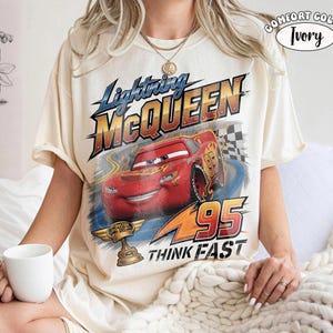 May include: Ivory-colored t-shirt featuring a graphic of Lightning McQueen from the Cars movie. The design includes the text "Lightning McQueen", "95", and "Think Fast". The shirt also has a Comfort Color label.