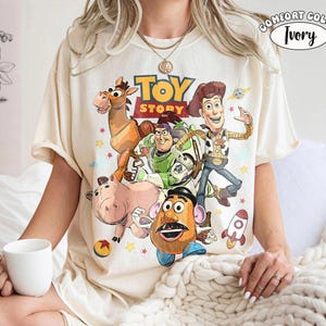 May include: Ivory-colored t-shirt featuring a colorful graphic of characters from the animated film "Toy Story." The design includes Woody, Buzz Lightyear, Mr. Potato Head, and other characters. The shirt has short sleeves and a crew neck.