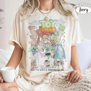 K&ouml;nnte beinhalten: Ein elfenbeinfarbenes Kurzarm-T-Shirt mit einem Grafikdruck von Toy Story-Figuren wie Woody, Buzz Lightyear, Jessie und Rex. Der Text lautet TOY STORY und YOU'VE GOT A FRIEND IN ME. Ein kleines kreisf&ouml;rmiges Etikett in der Ecke zeigt COMFORT COLOR IVORY.