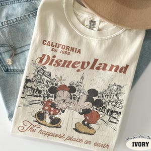May include: An ivory t-shirt featuring a graphic print of Mickey and Minnie Mouse in front of a castle. The text reads California Est. 1955 Disneyland and The happiest place on earth. The shirt has a Comfort Colors label at the inner neckline.