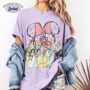 Op de afbeelding: Een lila Comfort Colors T-shirt met een afbeelding van Disney-prinsessen. Het ontwerp bevat een roze strik, een kasteel en vuurwerk. Het shirt heeft de tekst "Comfort Color Orchid" in een cirkelvormig ontwerp.