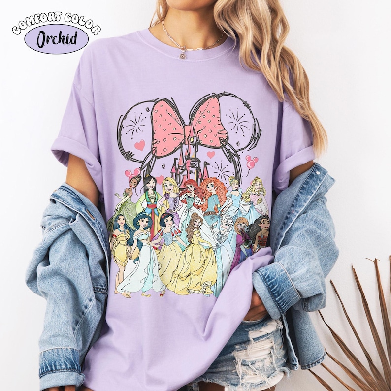 Op de afbeelding: Een lila Comfort Colors T-shirt met een afbeelding van Disney-prinsessen. Het ontwerp bevat een roze strik, een kasteel en vuurwerk. Het shirt heeft de tekst "Comfort Color Orchid" in een cirkelvormig ontwerp.