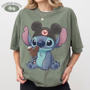 May include: A moss green t-shirt featuring a graphic of the character Stitch wearing a black ear hat with a red circular emblem. Stitch is holding a brown chocolate-covered ice cream bar shaped like mouse ears. The text Comfort Color Moss is visible.