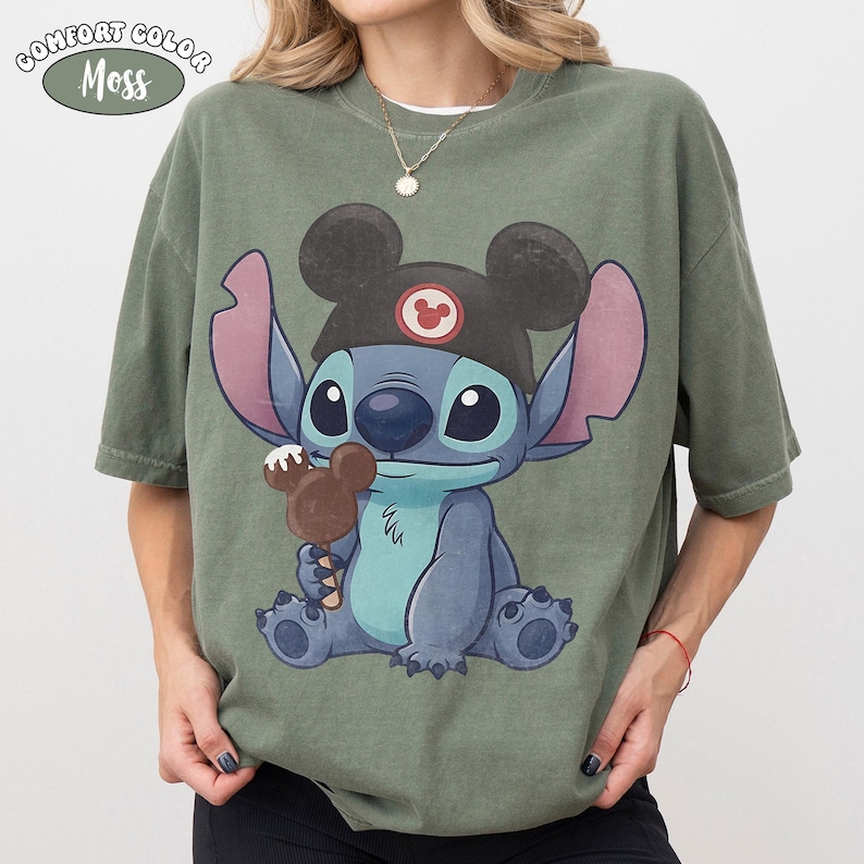May include: A moss green t-shirt featuring a graphic of the character Stitch wearing a black ear hat with a red circular emblem. Stitch is holding a brown chocolate-covered ice cream bar shaped like mouse ears. The text Comfort Color Moss is visible.