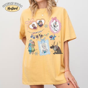 May include: Mustard-colored t-shirt featuring various characters and scenes from the animated film "Frozen." The design includes framed portraits of characters, snowflakes, and the word "FROZEN." The shirt has short sleeves and a relaxed fit.