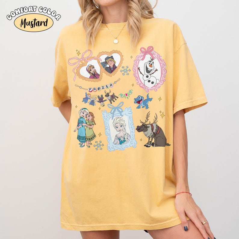 May include: Mustard-colored t-shirt featuring various characters and scenes from the animated film "Frozen." The design includes framed portraits of characters, snowflakes, and the word "FROZEN." The shirt has short sleeves and a relaxed fit.