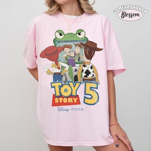 Pu&ograve; includere: T-shirt rosa Comfort Colors con grafica di Toy Story 5. Il design presenta Woody, Buzz Lightyear e Jessie. Il testo "TOY STORY 5" &egrave; in grandi lettere colorate. Il logo Disney Pixar &egrave; in basso.