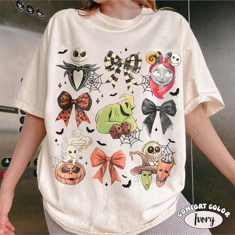 May include: An ivory short-sleeve t-shirt featuring a graphic print of characters and bows from The Nightmare Before Christmas. The design includes Jack Skellington, Sally, Oogie Boogie, Zero, and various bows, spiderwebs, and bats.
