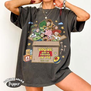 K&ouml;nnte beinhalten: Dunkelgraues T-Shirt mit einem Aufdruck von Toy Story-Charakteren in einer Kiste. Der Text "YOU'VE GOT A FRIEND IN ME SINCE 1995" ist darunter gedruckt. Auf dem Shirt steht auch "COMFORT COLOR Pepper" am unteren Rand.