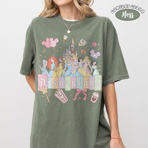 Puede incluir: Camiseta verde musgo con la palabra "Disneyland" en colores pastel, con ilustraciones de princesas Disney, un castillo e iconos del parque. Fabricada en material Comfort Colors.