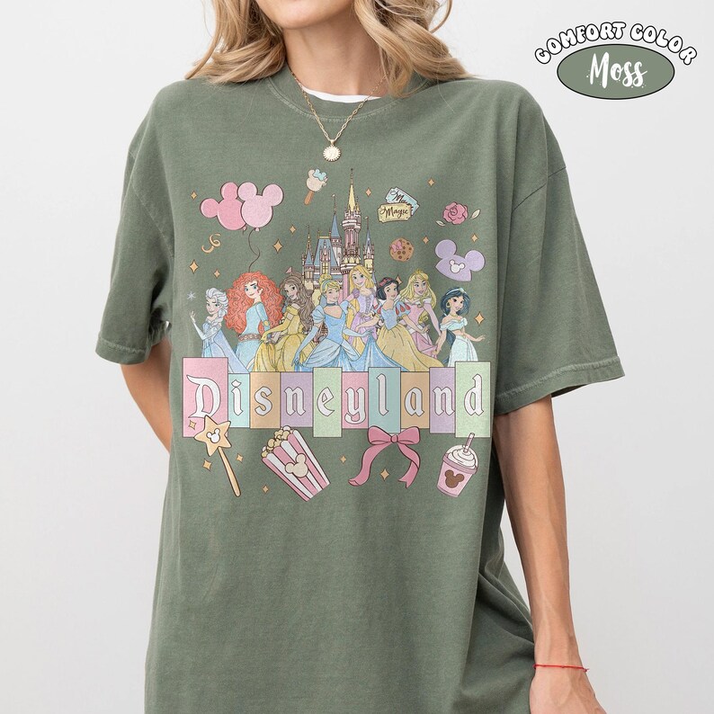 Puede incluir: Camiseta verde musgo con la palabra "Disneyland" en colores pastel, con ilustraciones de princesas Disney, un castillo e iconos del parque. Fabricada en material Comfort Colors.