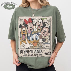 Puede incluir: Una camiseta verde musgo con un gr&aacute;fico rectangular de personajes de Disney como Mickey Mouse, Minnie Mouse, el Pato Donald, Goofy y Pluto. El texto dice Disneyland Where Dreams Come True 2026, con firmas de personajes y un borde vintage.