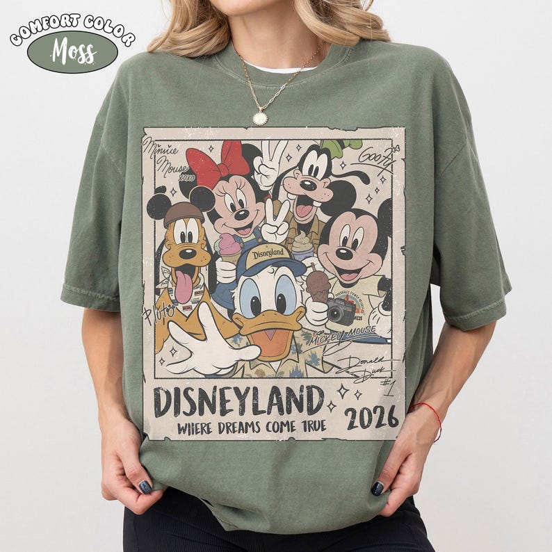 Puede incluir: Una camiseta verde musgo con un gr&aacute;fico rectangular de personajes de Disney como Mickey Mouse, Minnie Mouse, el Pato Donald, Goofy y Pluto. El texto dice Disneyland Where Dreams Come True 2026, con firmas de personajes y un borde vintage.