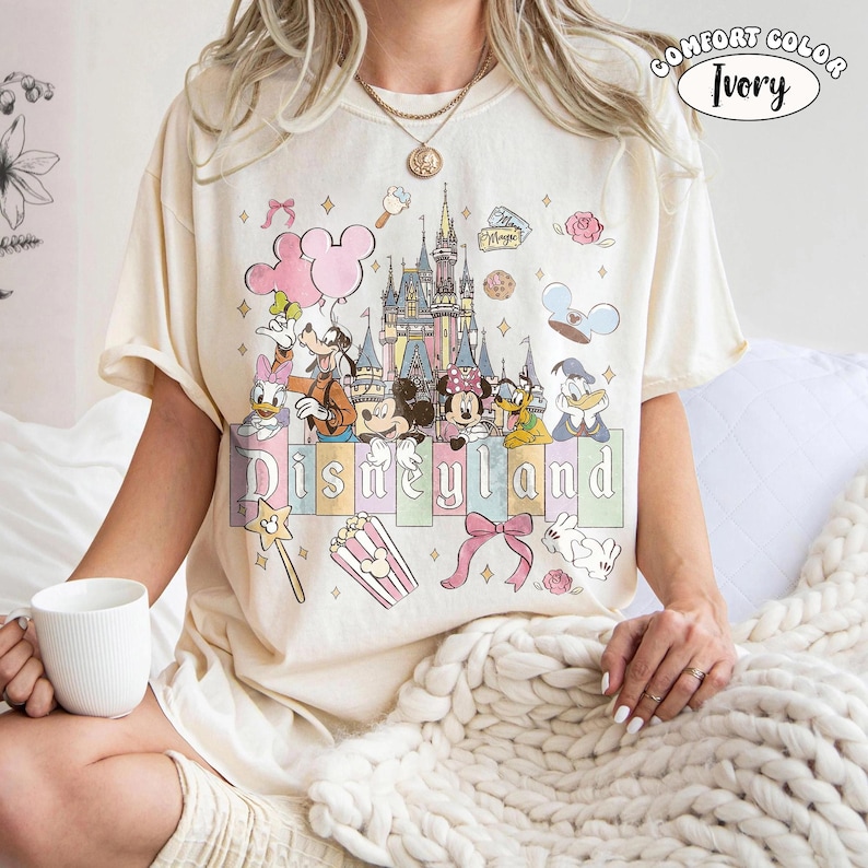 Peut inclure: T-shirt ivoire Comfort Colors avec un motif Disneyland. Le design comprend Mickey et Minnie Mouse, Donald Duck et un ch&acirc;teau. Le mot "Disneyland" est affich&eacute; en couleurs pastel, avec des ic&ocirc;nes th&eacute;matiques.