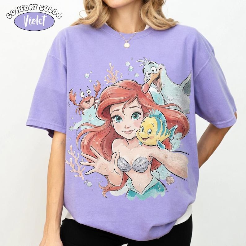 May include: Lilac Comfort Colors t-shirt with a vibrant graphic of Ariel, Flounder, Sebastian, and Scuttle from The Little Mermaid. The design is set against a lilac background. The text "Comfort Color" and "Violet" are in a circle at the top left.