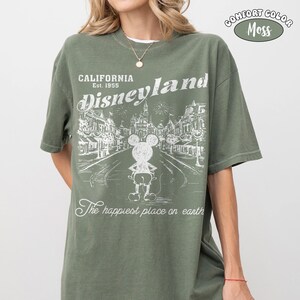 May include: Olive green t-shirt with a white graphic of Disneyland, featuring Mickey Mouse and the text "California Disneyland Est. 1955." The shirt also has the text "The happiest place on earth" and "Comfort Color Moss."