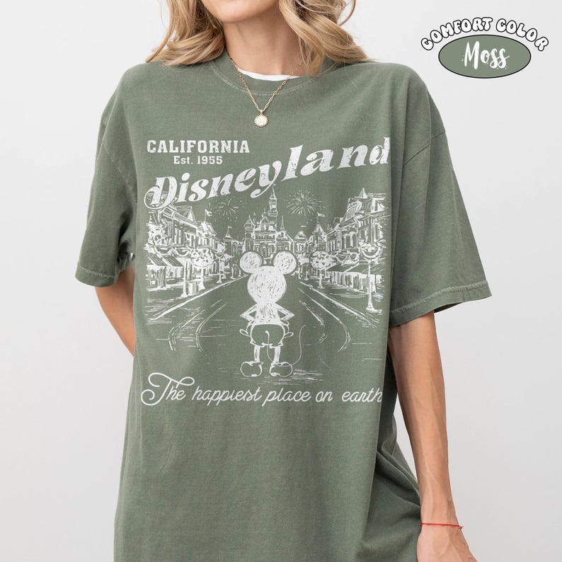 May include: Olive green t-shirt with a white graphic of Disneyland, featuring Mickey Mouse and the text "California Disneyland Est. 1955." The shirt also has the text "The happiest place on earth" and "Comfort Color Moss."