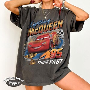 May include: Dark gray t-shirt featuring Lightning McQueen from the movie Cars. The design includes McQueen, the number 95, and the words "THINK FAST." The shirt also has the words "COMFORT COLOR Pepper" printed on it.