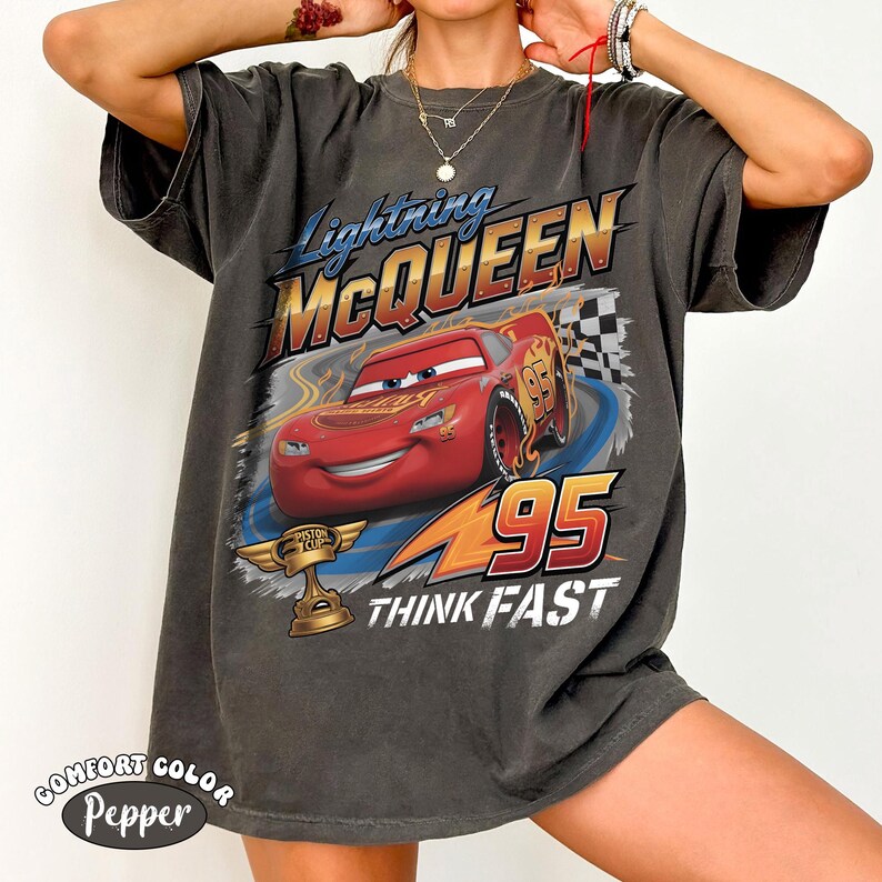 May include: Dark gray t-shirt featuring Lightning McQueen from the movie Cars. The design includes McQueen, the number 95, and the words "THINK FAST." The shirt also has the words "COMFORT COLOR Pepper" printed on it.