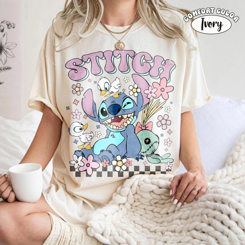 Puede incluir: Camiseta marfil Comfort Colors con un gr&aacute;fico de Stitch, patos y flores. La palabra "STITCH" est&aacute; en una fuente de burbujas. El dise&ntilde;o est&aacute; sobre un patr&oacute;n de cuadros blancos y negros. La camiseta es de manga corta.