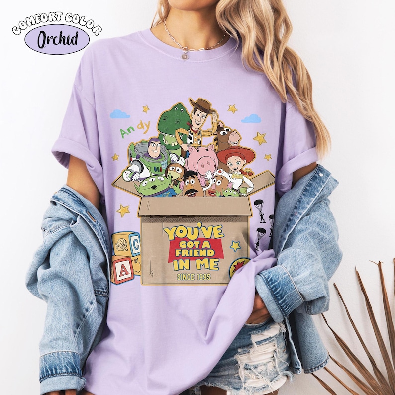 K&ouml;nnte beinhalten: Ein lila Comfort Colors T-Shirt mit einer Grafik von Toy Story-Charakteren. Das Design enth&auml;lt den Text "YOU'VE GOT A FRIEND IN ME SINCE 1995" und den Namen "Andy". Das Shirt hat kurze &Auml;rmel.