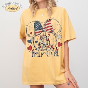 May include: Mustard-colored t-shirt featuring a graphic of a castle, Mickey Mouse ears, and a bow with an American flag design. The shirt has a vintage, distressed look with red, white, and blue hearts and fireworks.