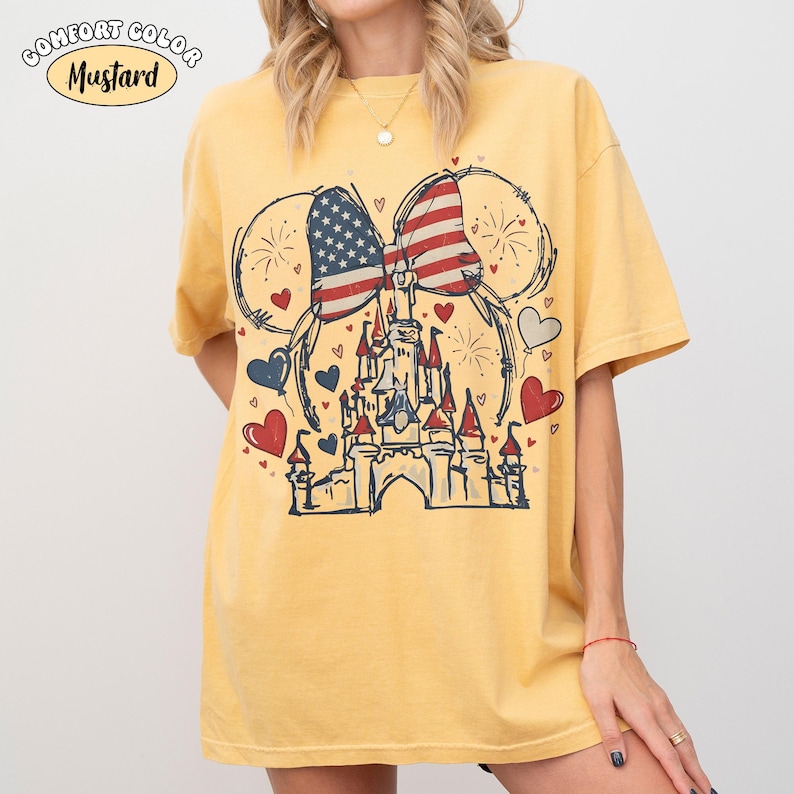 May include: Mustard-colored t-shirt featuring a graphic of a castle, Mickey Mouse ears, and a bow with an American flag design. The shirt has a vintage, distressed look with red, white, and blue hearts and fireworks.