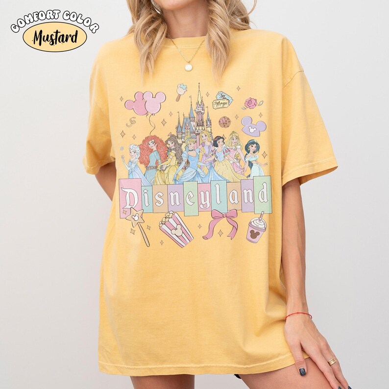 Puede incluir: Camiseta de color mostaza con un gr&aacute;fico de Disneyland. Presenta princesas de dibujos animados, un castillo y la palabra "Disneyland" en colores pastel. La camiseta tiene una etiqueta Comfort Color.
