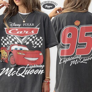 Może przedstawiać: Ciemnoszara koszulka z Lightning McQueen z filmu Auta Disneya i Pixara. Prz&oacute;d przedstawia postać z napisami "Cars" i "Lightning McQueen". Tył pokazuje duży napis "95" i "Lightning McQueen" w kolorze czerwonym.