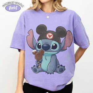 May include: A violet short-sleeve t-shirt featuring a graphic of the character Stitch wearing a black hat with mouse ears and holding a chocolate-covered mouse-shaped treat. The text COMFORT COLOR Violet is printed in the upper left corner.
