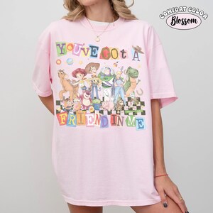 May include: Pink Comfort Colors t-shirt featuring a colorful graphic of characters and the phrase "YOU'VE GOT A FRIEND IN ME". The design includes a checkerboard pattern at the bottom. The shirt has short sleeves and a crew neck.