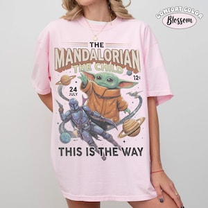 May include: A light pink t-shirt featuring a graphic print of The Mandalorian and The Child. The design includes planets, stars, and the text THE MANDALORIAN THE CHILD 24 JULY 12c THIS IS THE WAY. The shirt has short sleeves and a crew neckline.