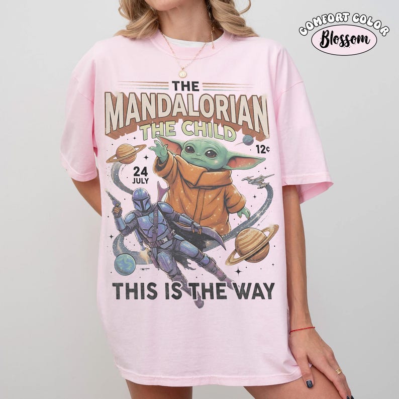 May include: A light pink t-shirt featuring a graphic print of The Mandalorian and The Child. The design includes planets, stars, and the text THE MANDALORIAN THE CHILD 24 JULY 12c THIS IS THE WAY. The shirt has short sleeves and a crew neckline.