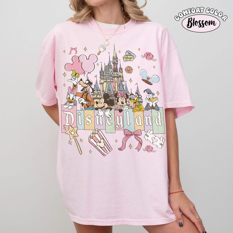 Peut inclure: T-shirt rose clair avec un motif color&eacute; de personnages Disney devant un ch&acirc;teau et le mot "Disneyland" en dessous. Le haut &agrave; droite porte l'inscription "Comfort Color Blossom" dans un cercle.
