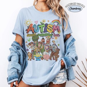 May include: A light blue t-shirt featuring a colourful graphic print with the text "AUTISM accept understand love" and various cartoon characters. The design includes a sun, a rainbow, and puzzle pieces, with a "COMFORT COLOR Chambray" label.