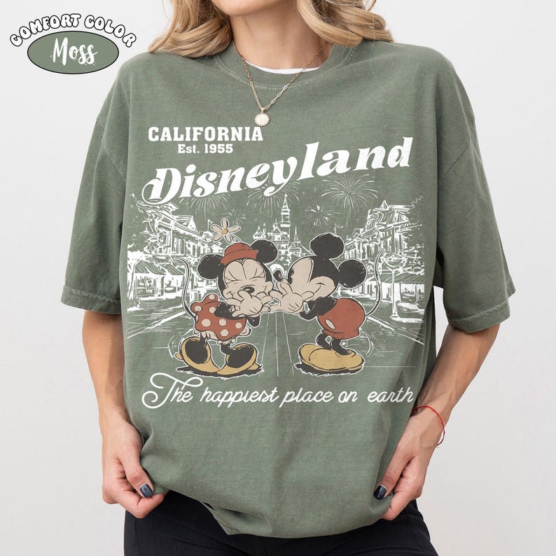 May include: A moss green short-sleeve t-shirt featuring a white graphic print of Mickey and Minnie Mouse. The text reads California Est. 1955 Disneyland The happiest place on earth. A small oval badge in the top left corner says Comfort Color Moss.
